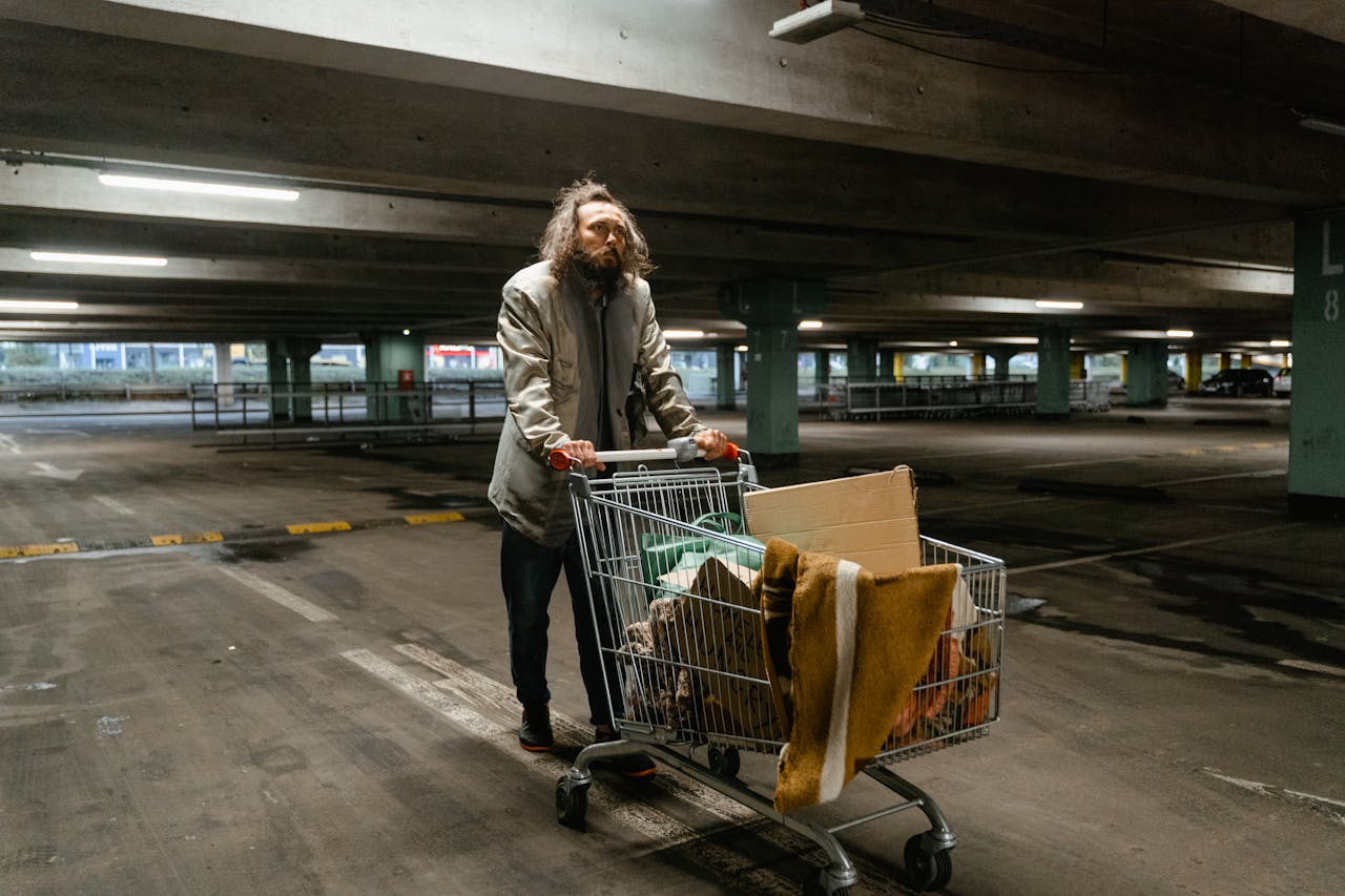 A homeless man pushing a shopping cart filled with belongings in a dimly lit parking garage.